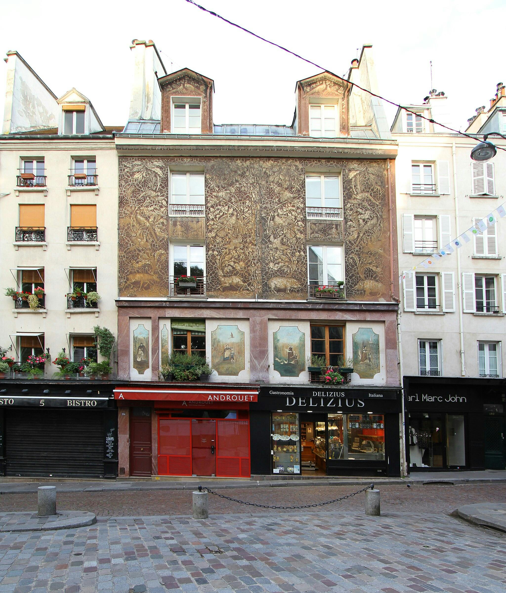 Facciata di un edificio europeo con opere d'arte dettagliate, piante sui balconi e negozi a livello della strada. Strada di ciottoli in primo piano.