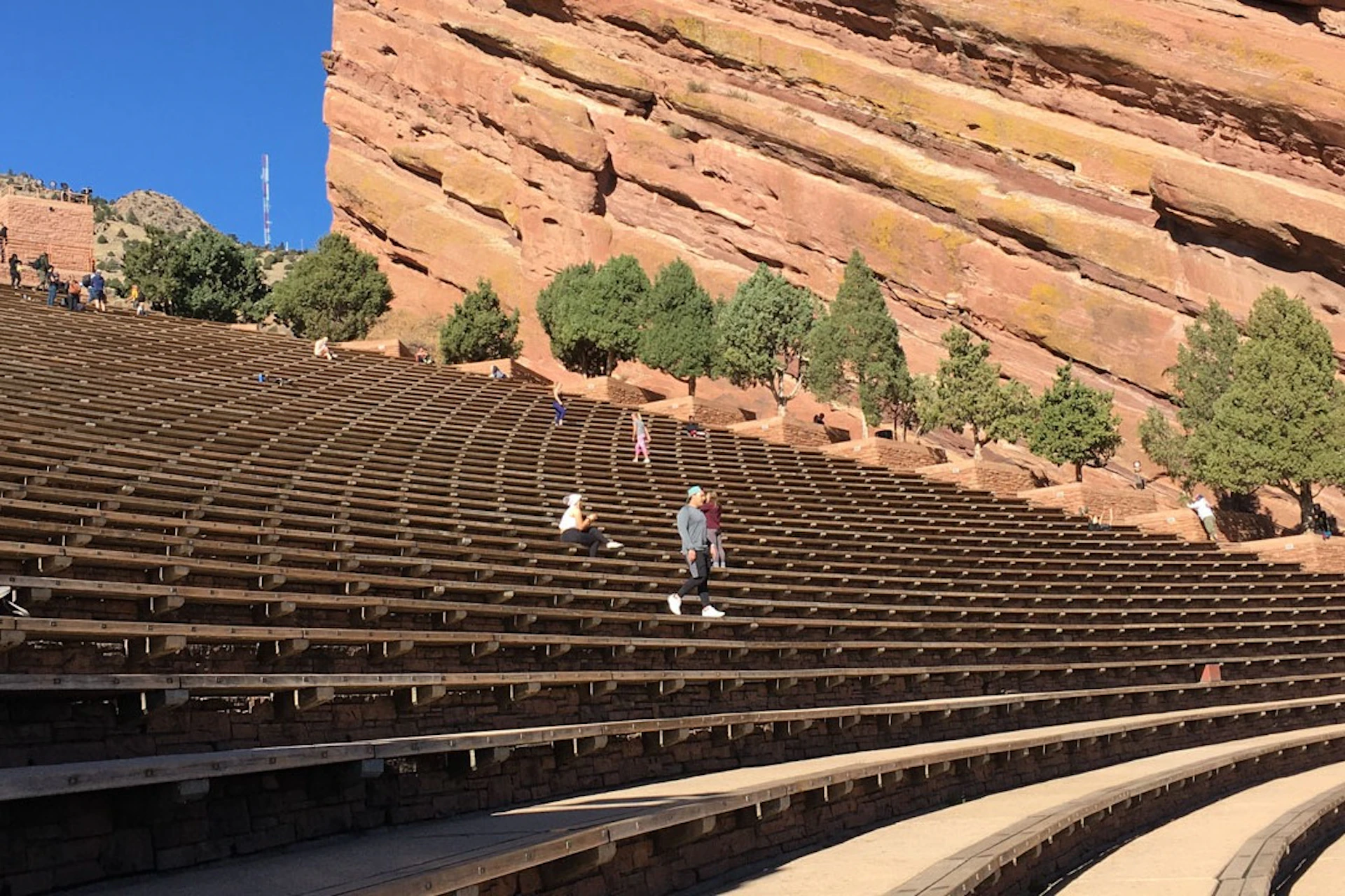 People walking and sitting on tiered stone seating with a backdrop of large red rock formations and sparse trees under a clear blue sky.