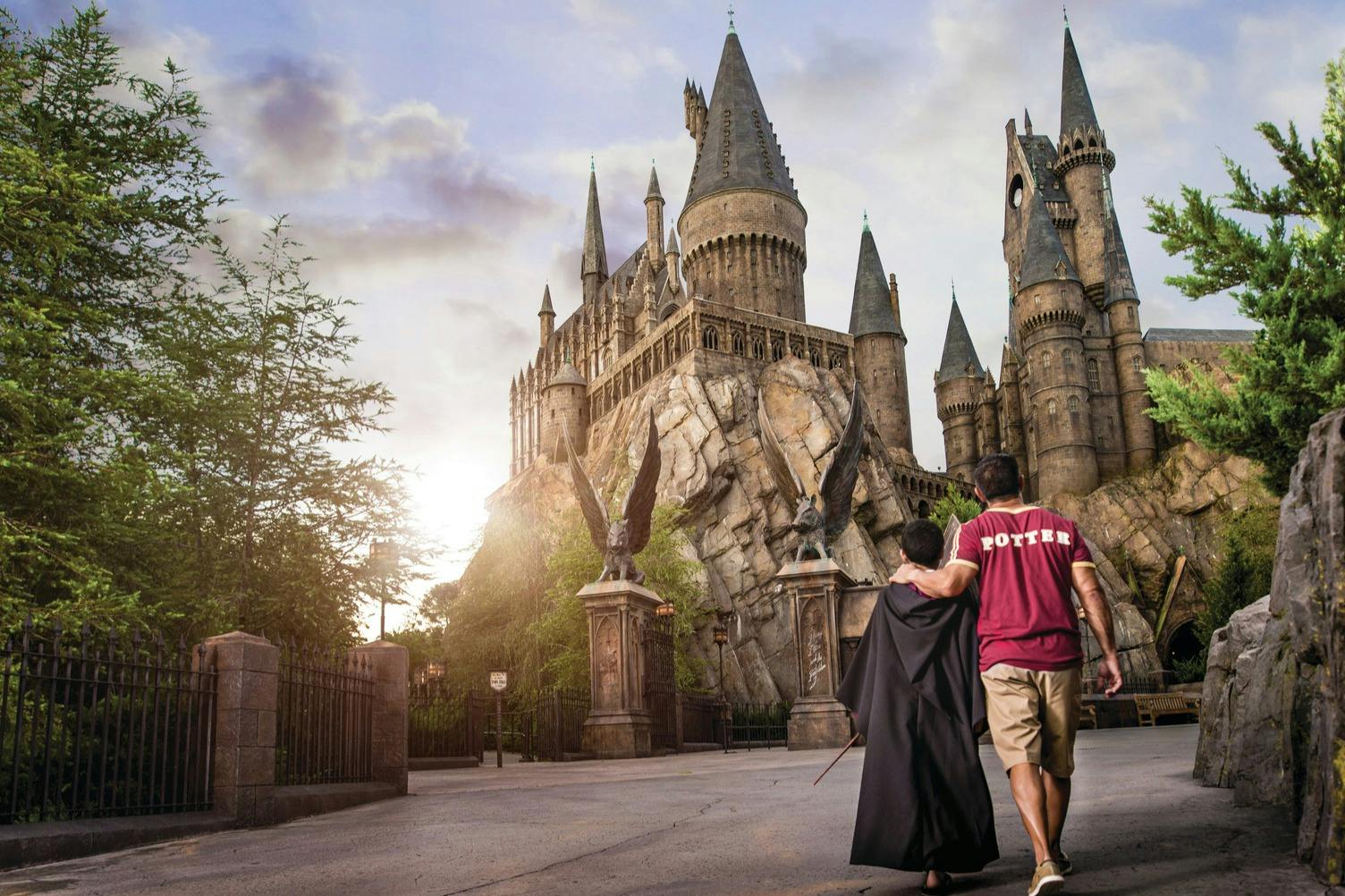 Due persone, una delle quali indossa una vestaglia di Harry Potter, camminano verso un grande castello che ricorda Hogwarts, con statue e verde intorno.