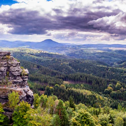 En naturskön vy över klippformationer med en naturstenbåge, omgiven av täta gröna skogar och under en delvis molnig himmel.