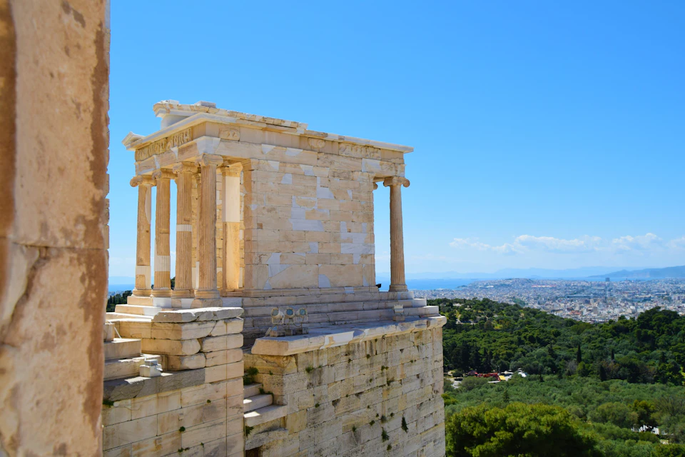 A small ancient Greek temple on a hill with a view of the city and greenery under a clear blue sky.