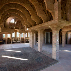 En rymlig interiör med tegelbågar, mönstrade mattor och naturligt ljus som strömmar genom fönster, vilket skapar en rustik atmosfär.