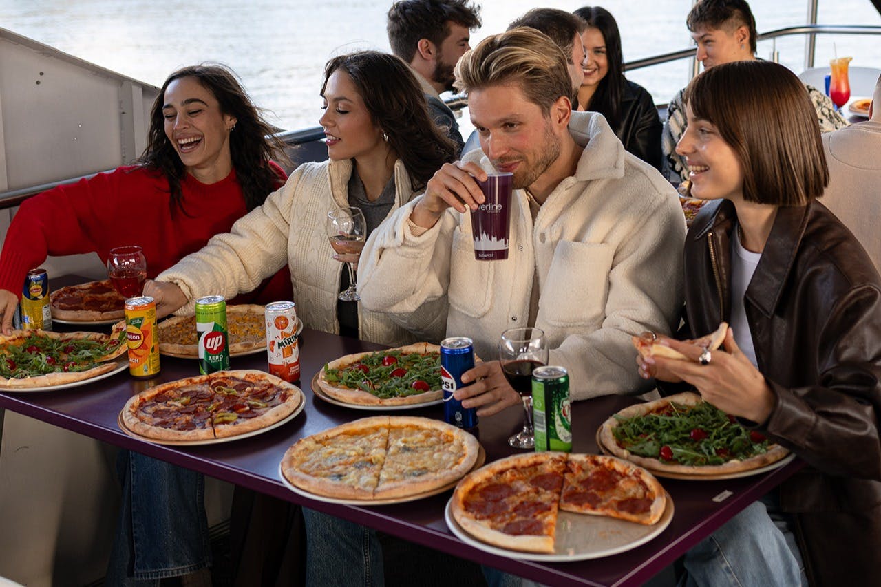 3人が屋外デッキのテーブルで様々なピザやドリンクを楽しんでいます。