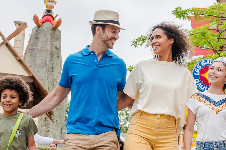 A family of four walks outdoors, smiling and interacting. A theme park attraction and greenery are visible in the background.