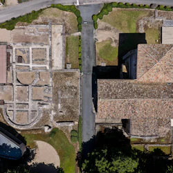 Flygfoto över forntida stenruiner och intilliggande byggnader med tegeltak, omgivna av vägar och grönområden.