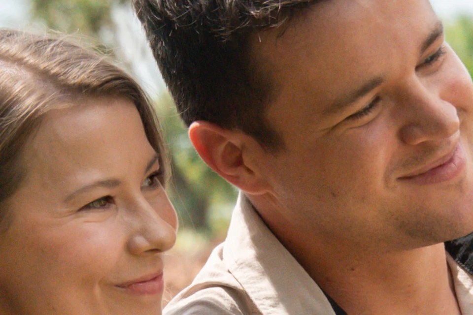 Two people outdoors, one holding a white cockatoo bird. Both are smiling, surrounded by greenery under a bright sky.