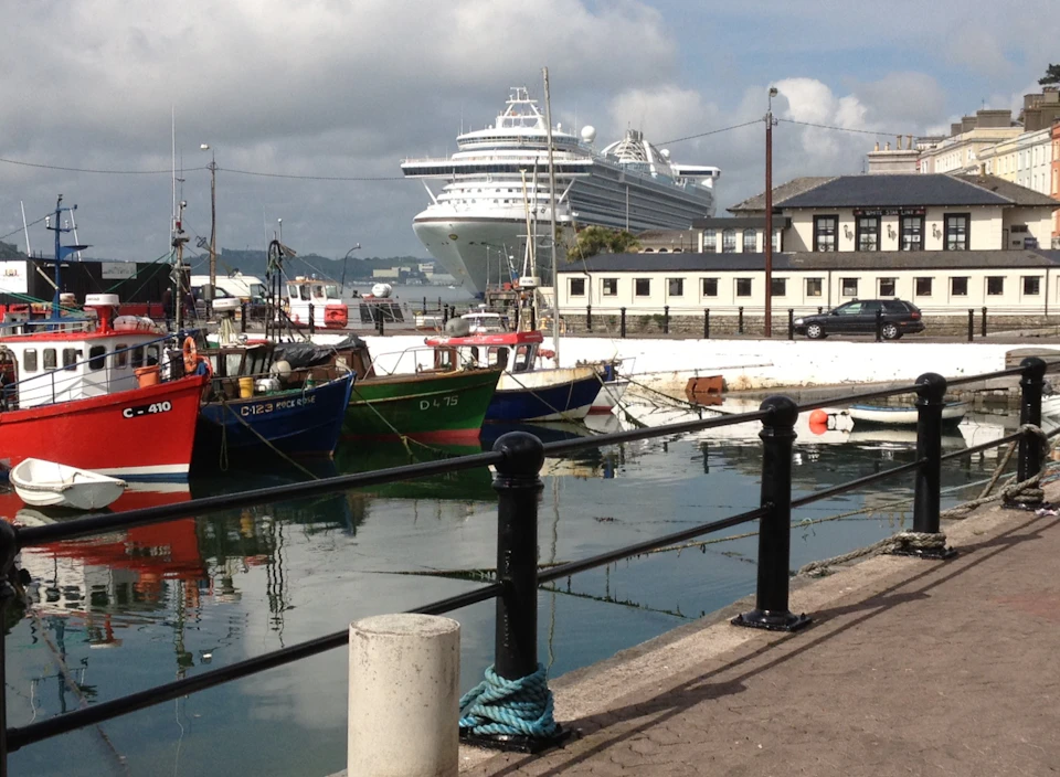 Fishing boats docked at a harbor with a large cruise ship and buildings in the background.