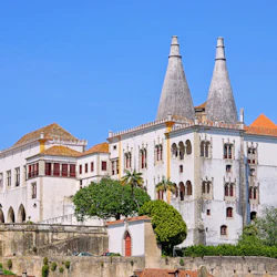 Historiskt vitt palats med koniska skorstenar, rödklädda fönster och palmer, mot en klarblå himmel.