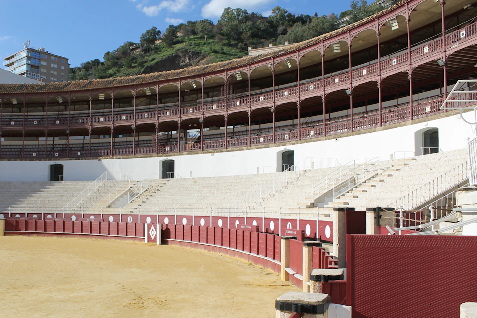 Empty bullring with red and white barricades, stone seating, and arched covered stands against a backdrop of green hillside.