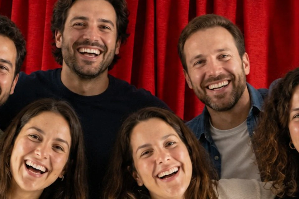 Six smiling people stand in front of a red curtain under a sign that reads 'El Felicitador' in black letters.