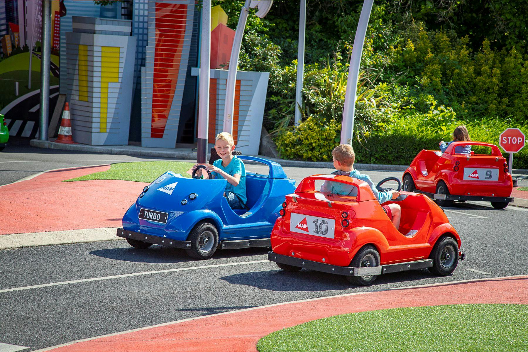 Two children driving mini cars, one blue and one red, on a small track in an outdoor amusement area with greenery.