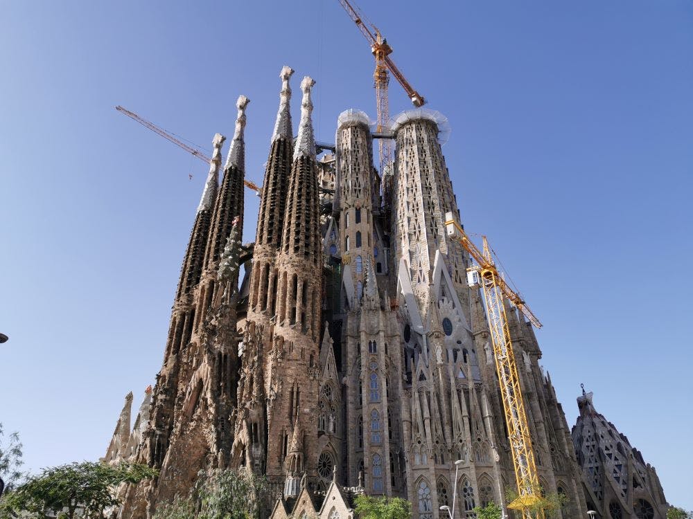 A large, ornate cathedral under construction with tall spires and cranes set against a clear blue sky.