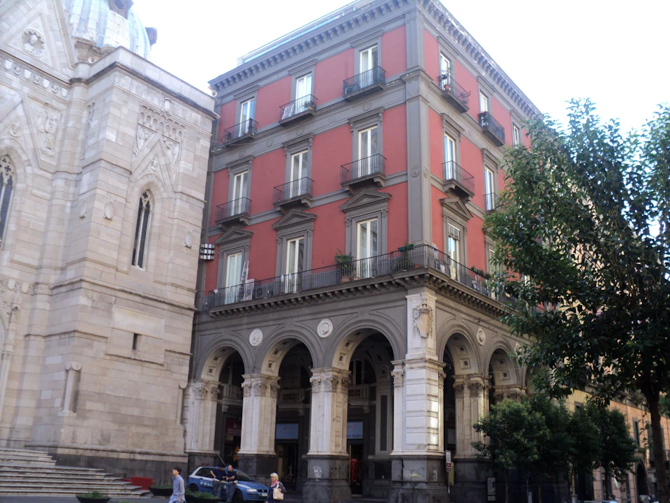 A four-story red building with arches on the ground level, located beside a stone structure with arched windows.