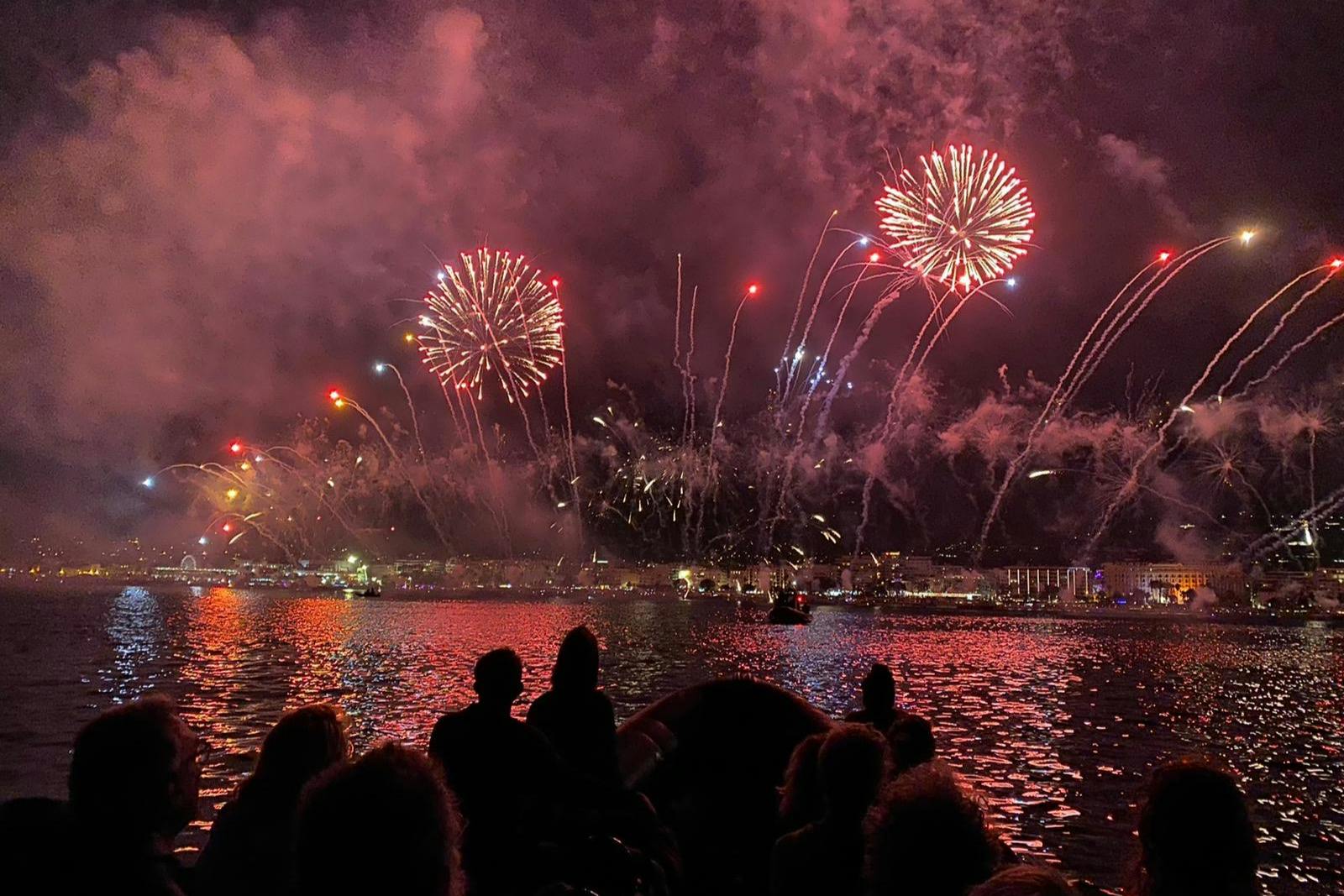 Silhouetted people watching colorful fireworks burst over a city waterfront at night.