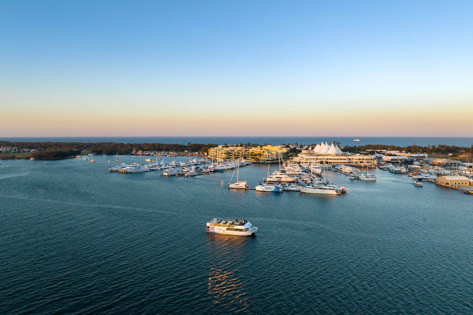 Aerial view of a marina with numerous boats docked, a ferry sailing in open water, and buildings on the shoreline at sunset.