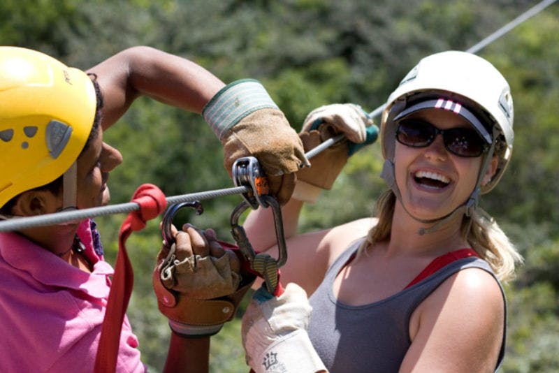 Personne portant un casque et des lunettes de soleil, souriant tout en se tenant à une tyrolienne avec des gants, en extérieur avec de la verdure en arrière-plan.