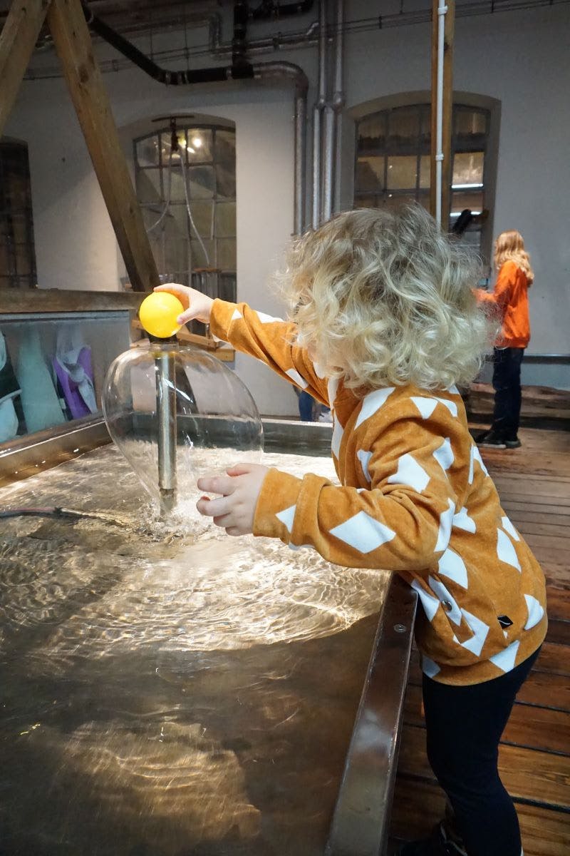 Ett barn med lockigt hår leker med ett vattenbord med hjälp av en gul boll. Ett annat barn i en orange skjorta syns i bakgrunden.