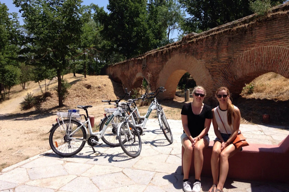 Two women sit on a bench near bicycles with a historic brick arch bridge in the background on a sunny day.