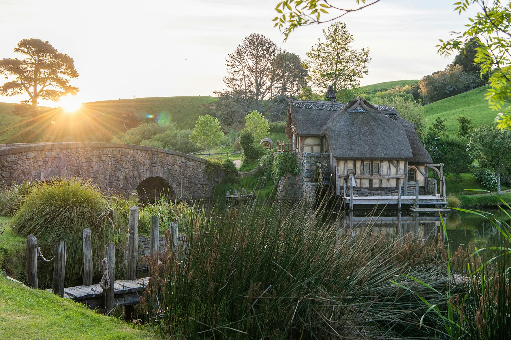A rustic stone bridge and a thatched cottage beside a small pond surrounded by lush greenery at sunset.