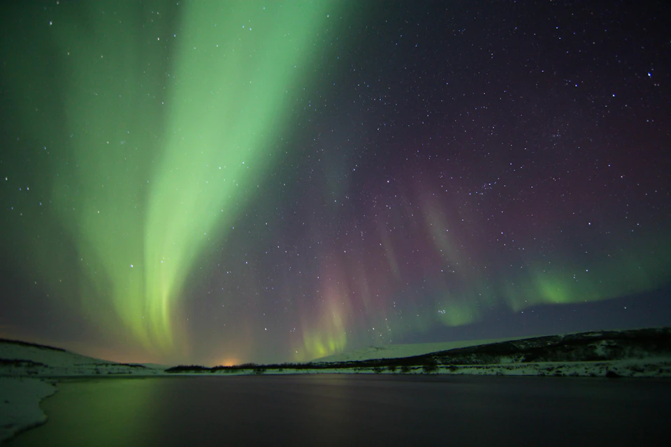 Aurora borealis with green and purple lights in the night sky over a calm body of water and silhouetted landscape.