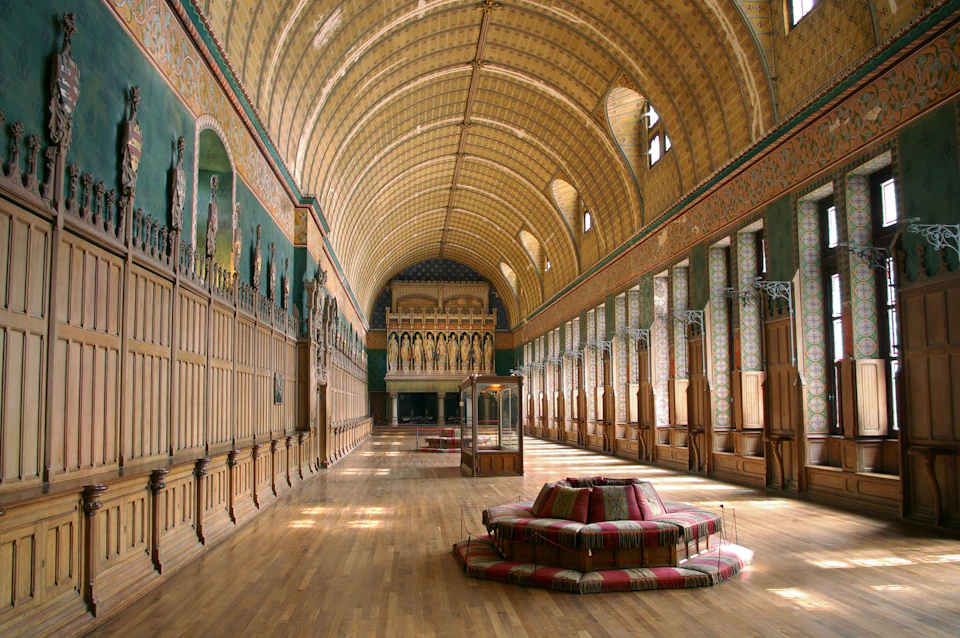 A grand hall with a vaulted ceiling, ornate wall decorations, wooden flooring, tall arched windows, and a central cushioned seating area.