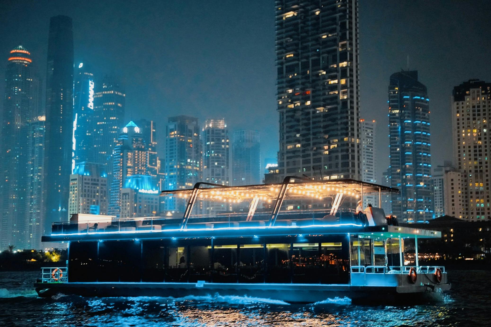 En modern båt med glödande ljus seglar på vatten på natten, med en bakgrund av upplysta skyskrapor.