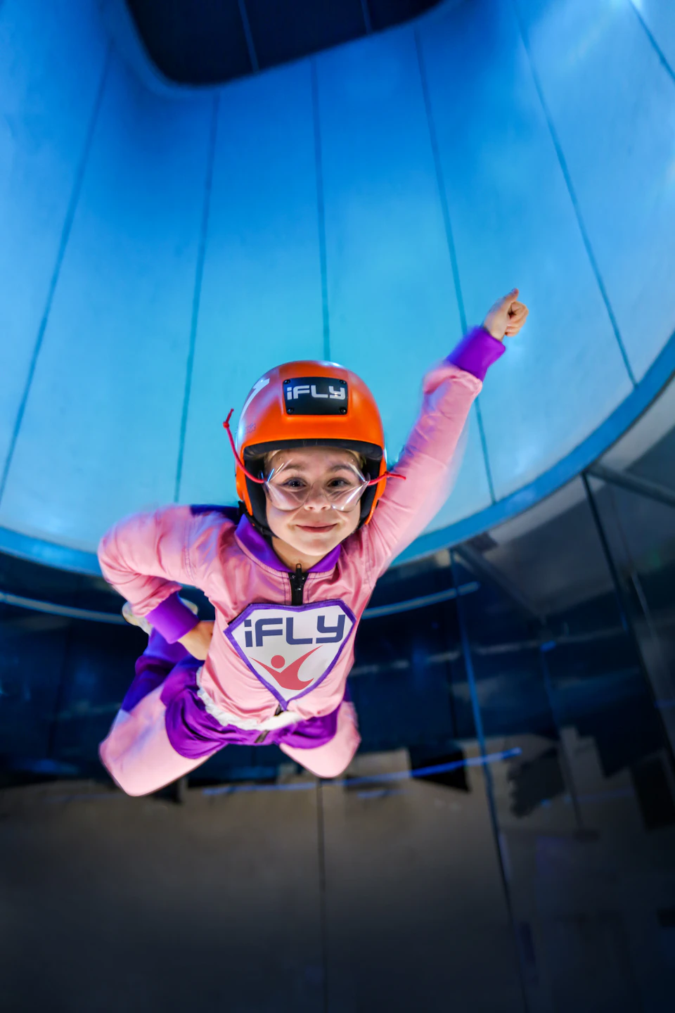 Child in pink and purple flight suit and orange helmet is indoor skydiving in a vertical wind tunnel.