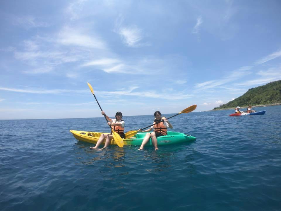 Two people wearing life jackets paddle kayaks on a calm blue sea under a bright sky with scattered clouds.
