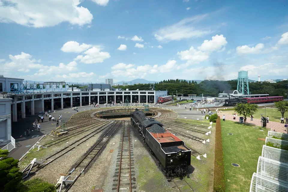 A roundhouse with a turntable and various railway tracks, featuring a black steam locomotive and people walking nearby.