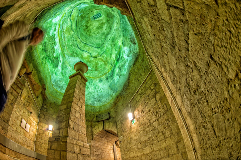 A historic stone chamber with a green-lit ceiling and an arched entrance, featuring a central pillar and wall-mounted lights.