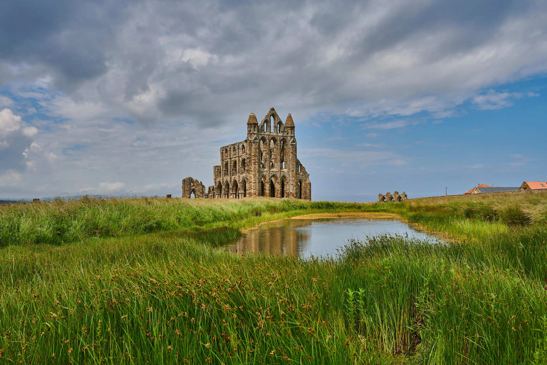 Ruins of a historic stone abbey surrounded by green grass and a small pond under a partly cloudy sky.
