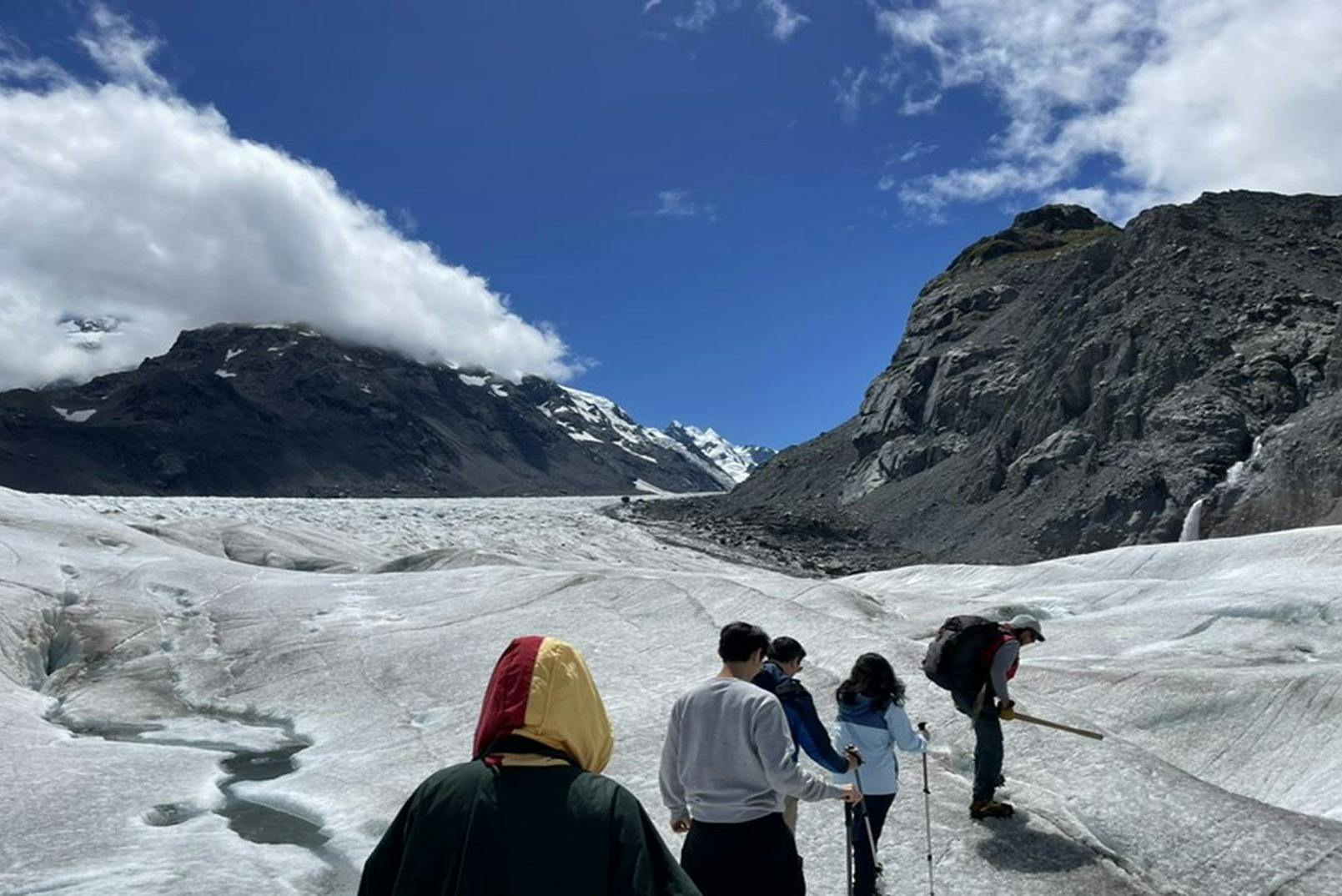A group of people walking on a glacier with snowy mountains and rocky peaks in the background under a clear blue sky.