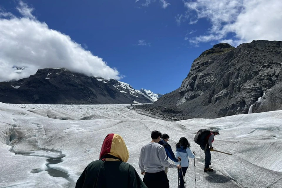 A group of people walking on a glacier with snowy mountains and rocky peaks in the background under a clear blue sky.