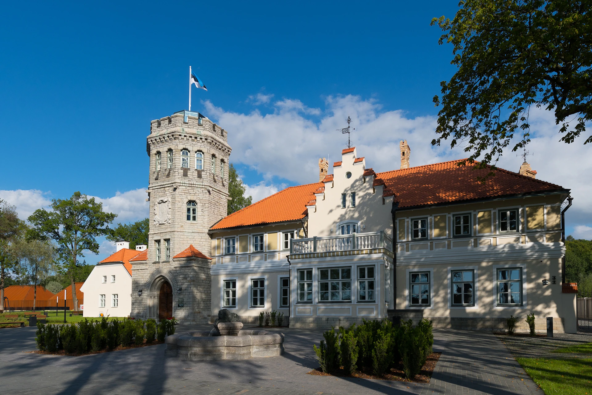 Historic building with a stone tower and an orange-tiled roof beneath a blue sky, featuring a flag atop the tower and well-kept shrubs.