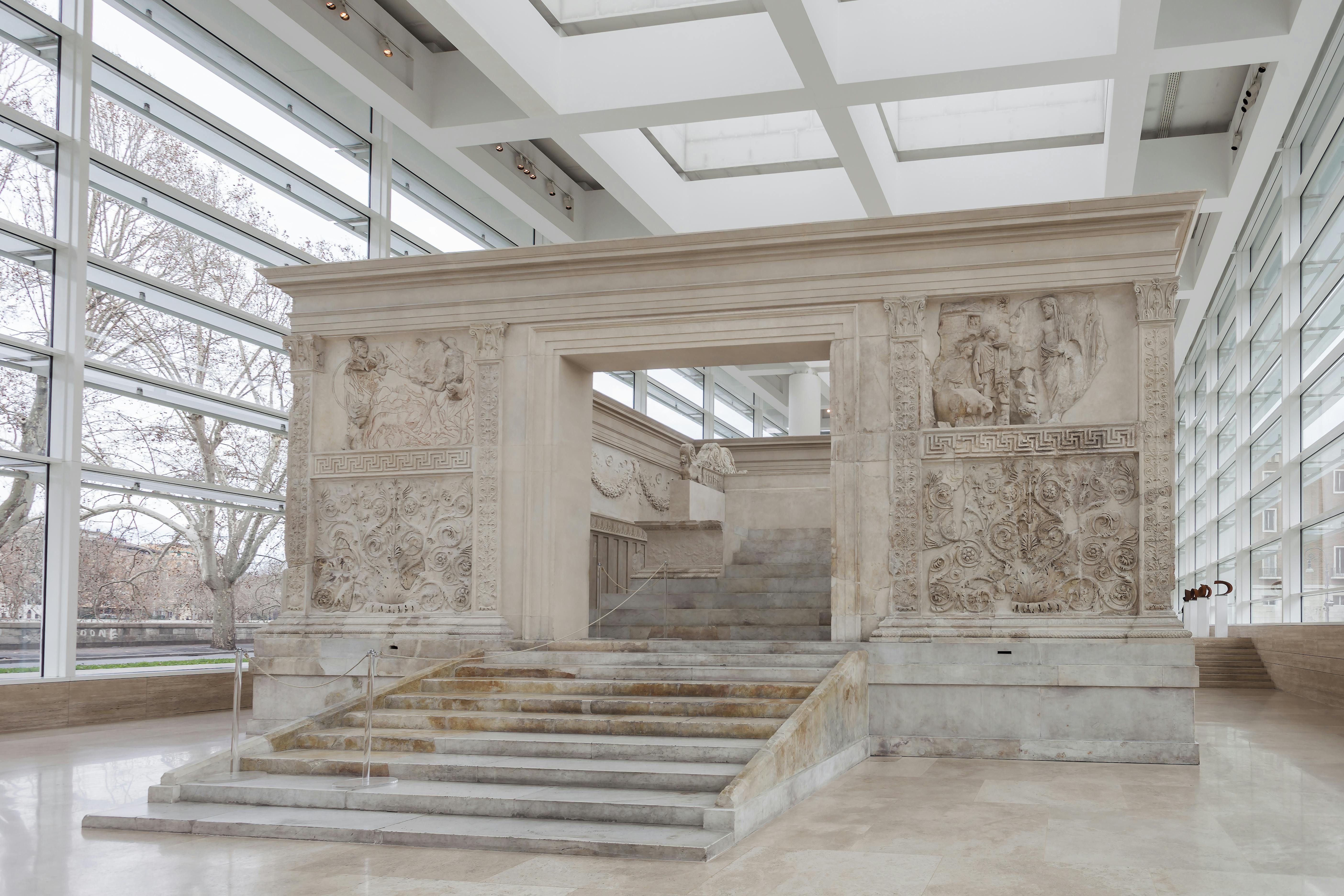 A marble structure with intricate carvings, positioned inside a modern glass-ceilinged room, with a wide staircase leading to it.