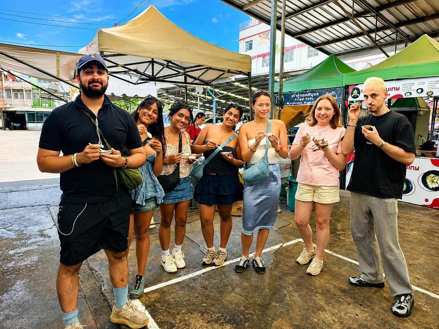 A group of people standing together outdoors at a market, each holding small items and smiling, with tents in the background.