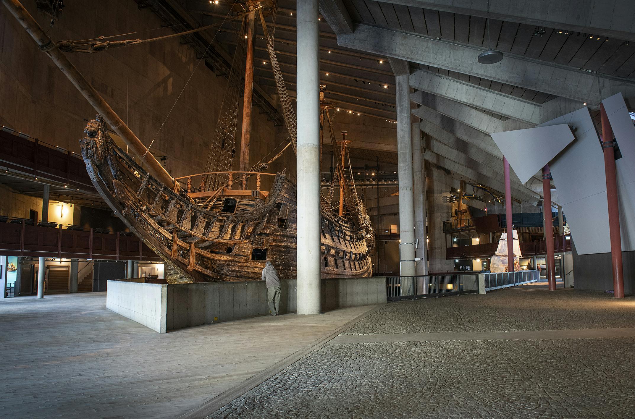 A large, historical wooden ship displayed indoors. A person in a gray jacket observes the ship in the spacious, modern museum.