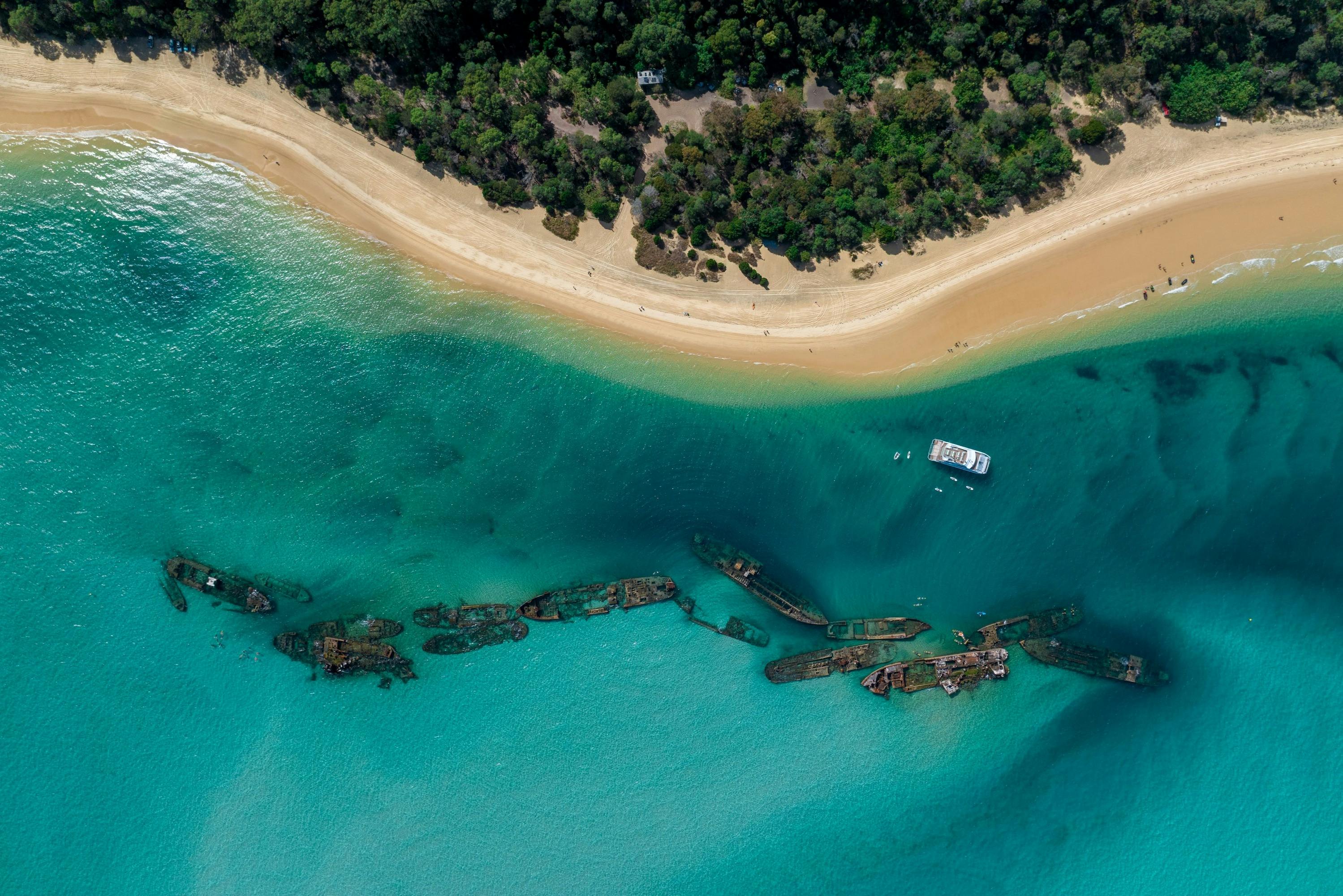 Luchtfoto van turquoise water, een zandstrand, groen bos, een witte boot en gedeeltelijk ondergedompelde scheepswrakken.