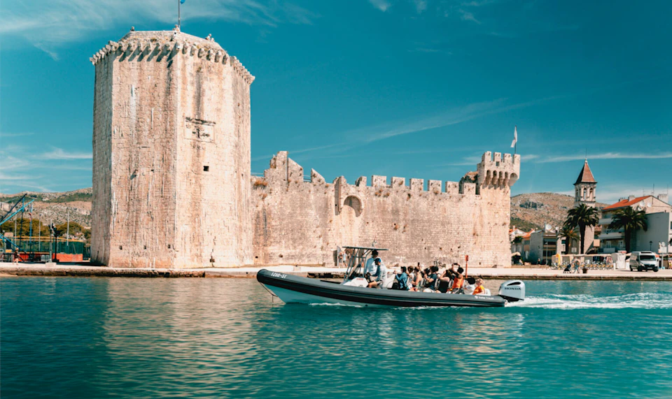 A small boat with people passes by a historic stone fortress on a sunny day, with clear blue water and sky in the background.