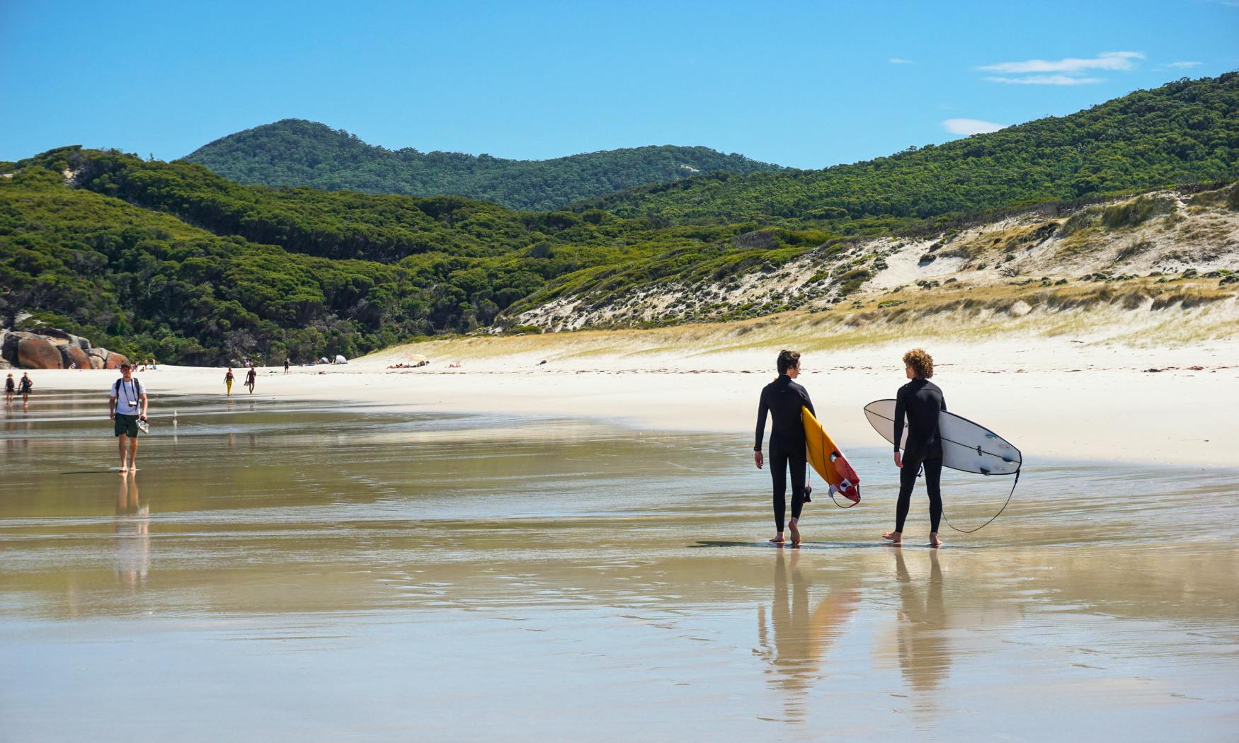 Δύο σέρφερ που κρατούν σανίδες περπατούν κατά μήκος μιας αμμώδους παραλίας προς τον ωκεανό. Στο βάθος διακρίνονται καταπράσινοι λόφοι.