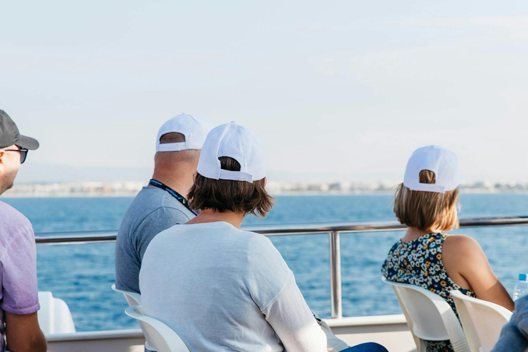 Três pessoas com bonés brancos sentam-se num barco, de frente para o mar, sob um céu limpo.