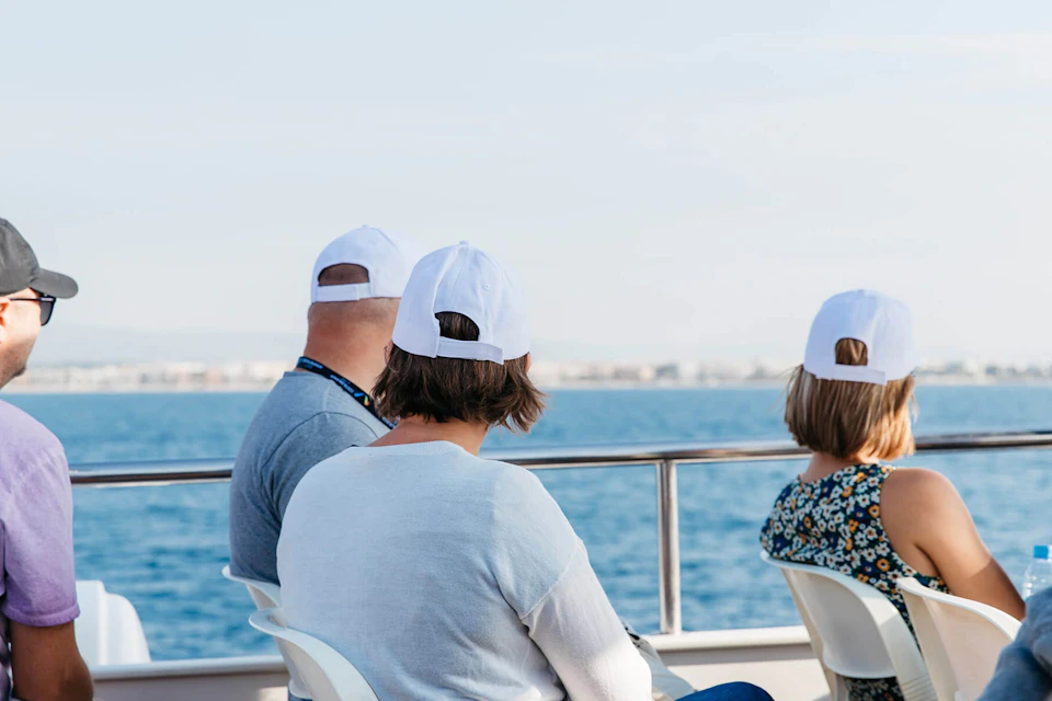 Three people wearing white caps sit on a boat, facing the sea under a clear sky.