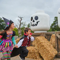 Two children in Halloween costumes run by large LEGO skull decorations and hay bales, with trees in the background.