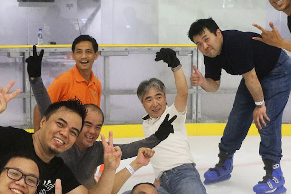 A group of men posing on an indoor ice-skating rink, some standing and others sitting on the ice, smiling and gesturing.