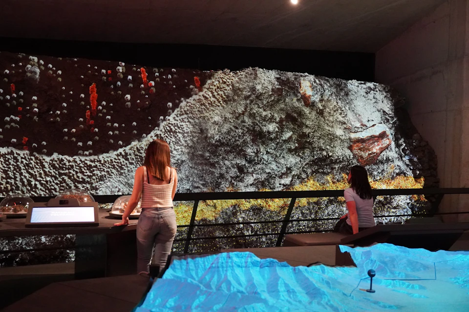 Two women observe a large geological display on a wall with a textured, mountainous landscape. A blue terrain model is in the foreground.