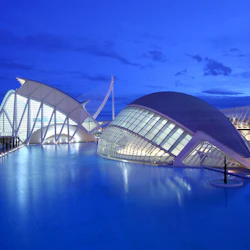 Futuristiska vita byggnader med krökta och kantiga former omgivna av vatten, upplysta mot en skymningsblå himmel.