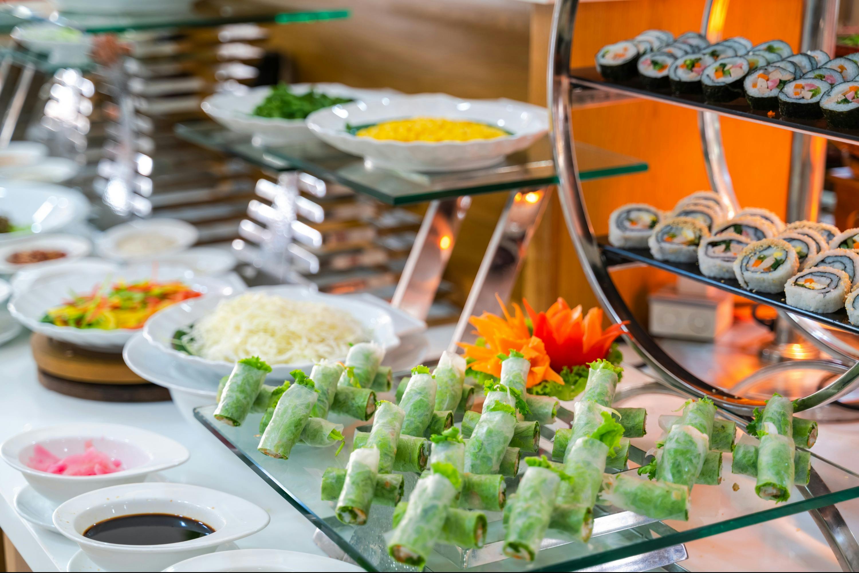 A buffet display featuring spring rolls, sushi, and various garnishes on glass shelves with a decorative arrangement.