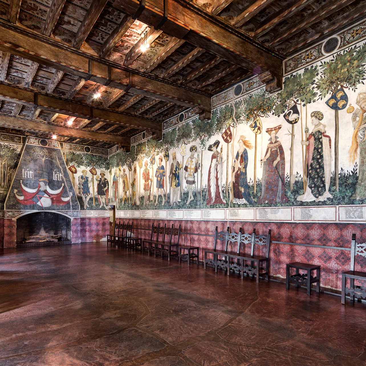 A historic hall with wooden beams, murals of medieval figures on the walls, a large fireplace, and rows of wooden chairs.
