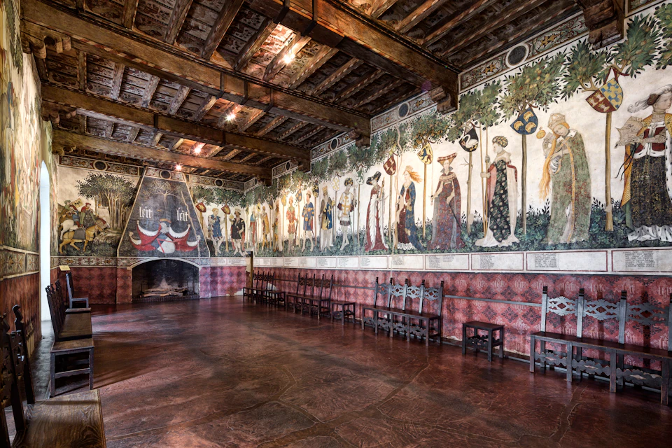 A historic hall with wooden beams, murals of medieval figures on the walls, a large fireplace, and rows of wooden chairs.