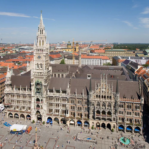 Luftaufnahme eines Gebäudes im gotischen Stil mit einem hohen Uhrenturm, umgeben von anderen Gebäuden und einem belebten Platz mit Menschen.